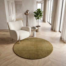 Beige round Hand Woven Loom Lori wool rug styled beneath a cream armchair in a bright modern interior with warm wooden flooring.